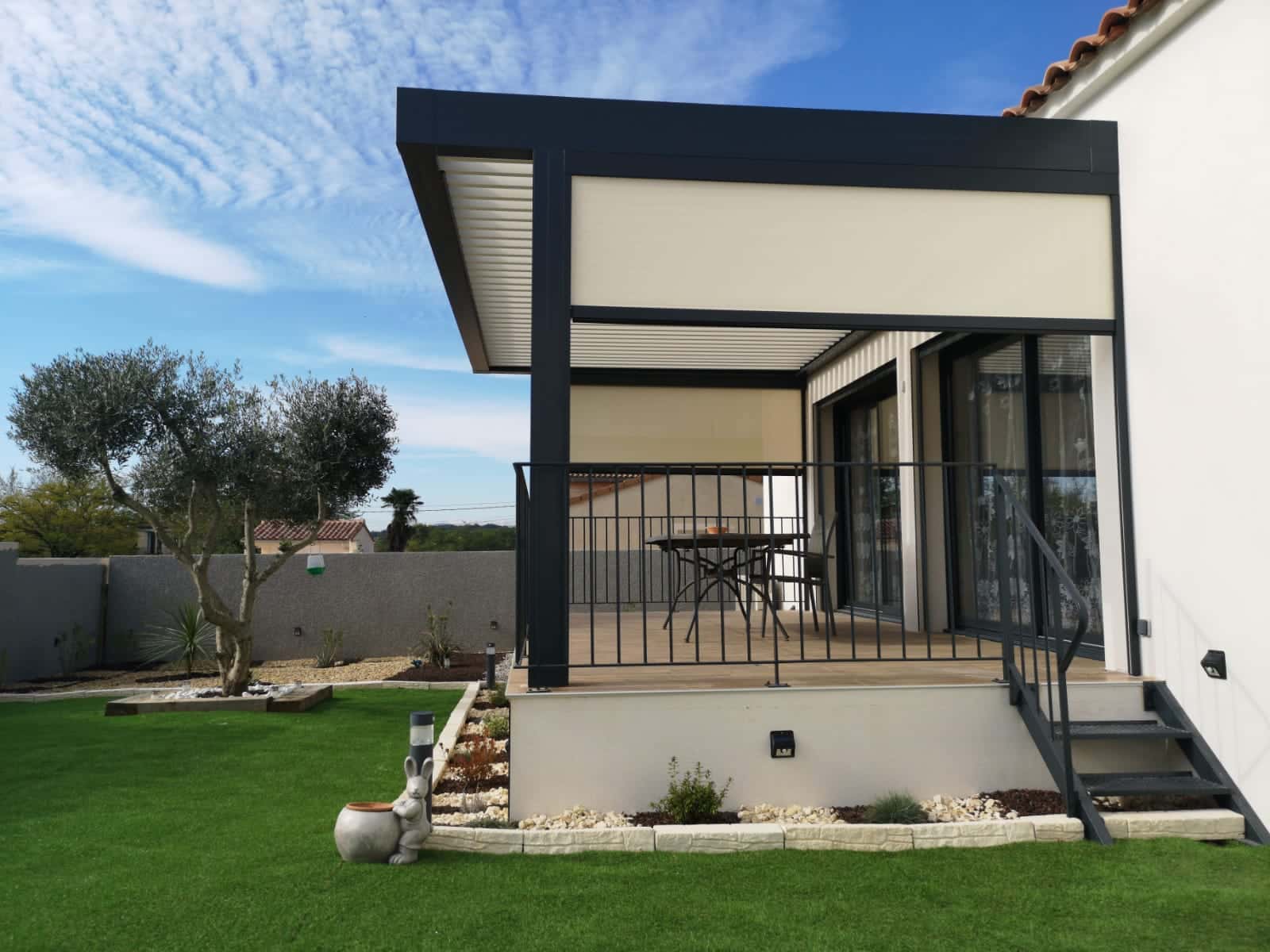 Pergola bioclimatique anthracite adossée une façade de maison, avec une gazon synthétique en premier plan.